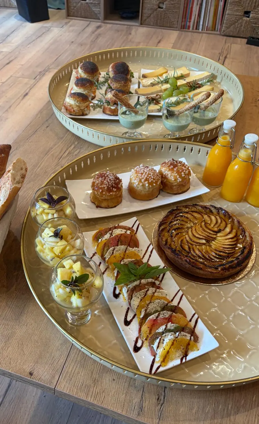 Le brunch dressé sur une table en bois : buns et fromages, brioches, tarte aux pommes, tomates et mozzarella, salades d’ananas, baguettes et jus d’orange pressé