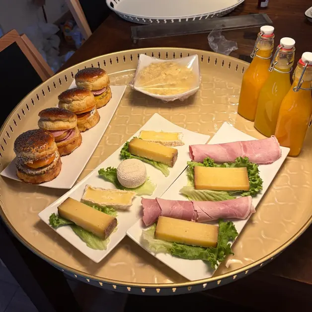 Un plateau doré avec des fromages et du jambon sur un lit de salade, des buns garnis, des œufs brouillés et trois bouteilles de jus d’orange pressé