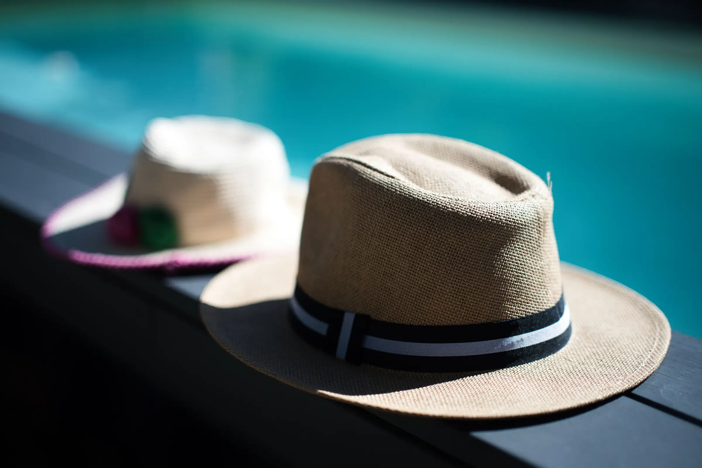 Deux chapeaux de paille posés sur la margelle de la piscine chauffée du Chalet Normand, l’eau turquoise derrière