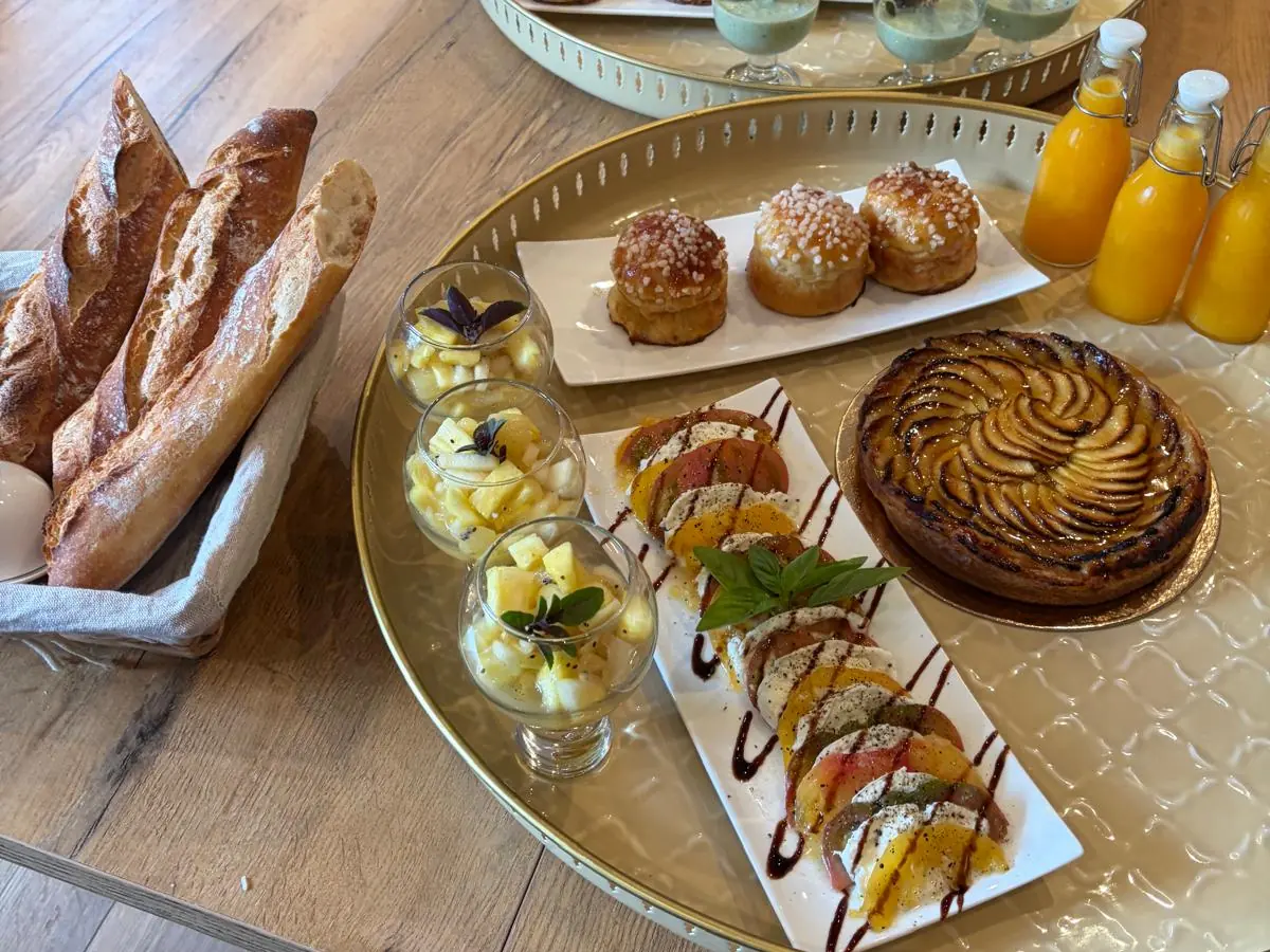 Les plateaux du matin dressés sur une table en bois : brioches au sucre, tarte aux pommes, tomates et mozzarella, salades d’ananas, baguettes et jus d’orange pressé