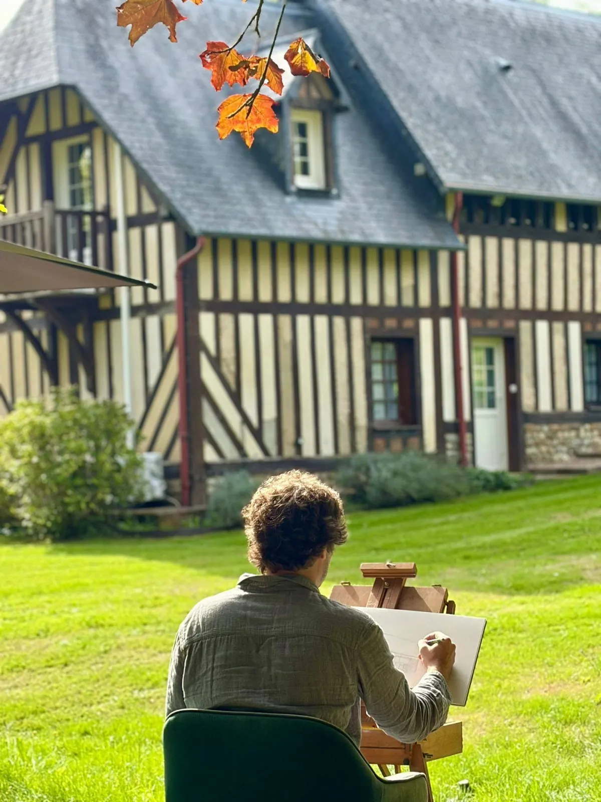 Cyp, de dos, dessine à son chevalet sur la pelouse, face à Reinette Grise, au Hameau des Prés-Verts, sous des feuilles d’automne