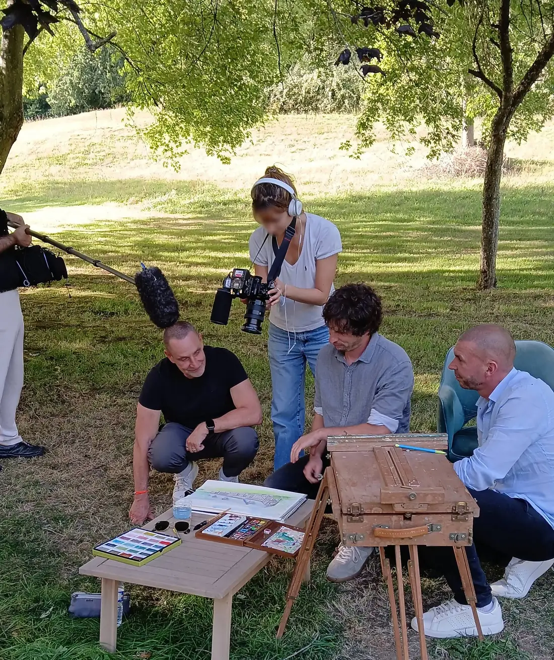 Le tournage au Hameau des Prés-Verts : Stéphane, accroupi, et Loïc, assis, penchés avec Cyp sur son aquarelle ; une cadreuse de France 2 filme au-dessus d’eux, une perche de son entre dans le cadre